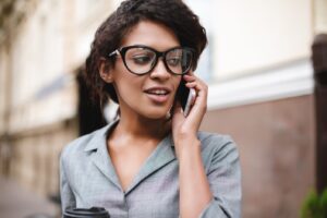 Beautiful African American girl in glasses standing on street and talking on her cellphone. Portrait of lady with dark curly hair in gray dress standing and dreamily looking aside on street